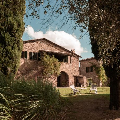 Outdoor terrace at Colle ai Lecci agriturismo, Chianti, with a linen-draped table set for coffee against a centuries-old stone and brick farmhouse facade with olive-green shutters and hydrangeas