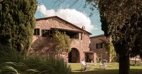 Outdoor terrace at Colle ai Lecci agriturismo, Chianti, with a linen-draped table set for coffee against a centuries-old stone and brick farmhouse facade with olive-green shutters and hydrangeas