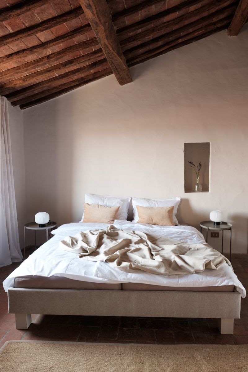 Serene boutique bedroom at Colle ai Lecci, Tuscany, with original restored timber beams, terracotta tile floors, linen bedding in natural tones and globe bedside lamps, designed by architect Nina Mair