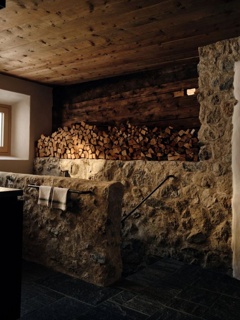 The stone-walled cellar at the Sonnenhof, split logs stacked the length of a rough natural stone wall uncovered and re-laid by hand, timber ceiling boards above and a small deep-set window.