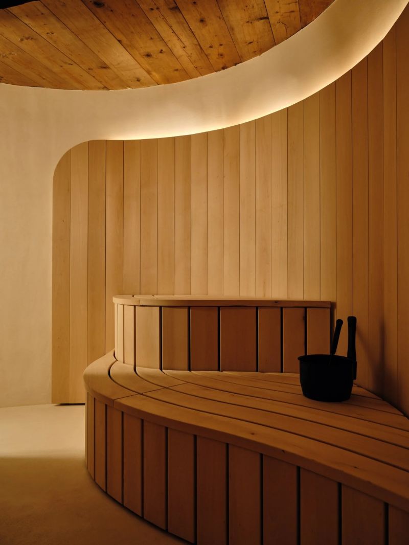The Finnish sauna by Shinichiro Ogata at the Sonnenhof, a curved slatted bench in blond timber against a sweeping limewash wall lit by a concealed cove, aged boards overhead.