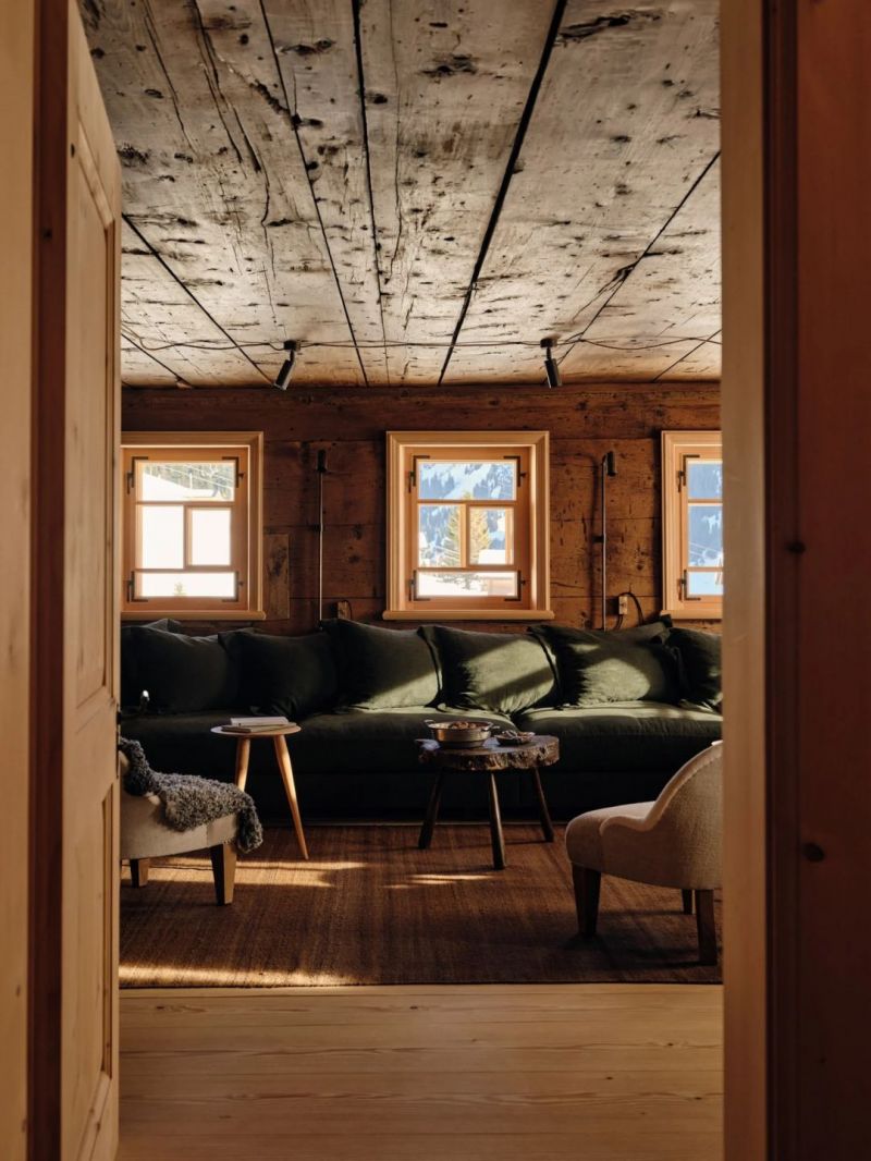 The lounge at the Sonnenhof seen through an open timber door, a long forest green banquette beneath three small windows, pale weathered ceiling boards and dark beams, sheepskin over a cream armchair.