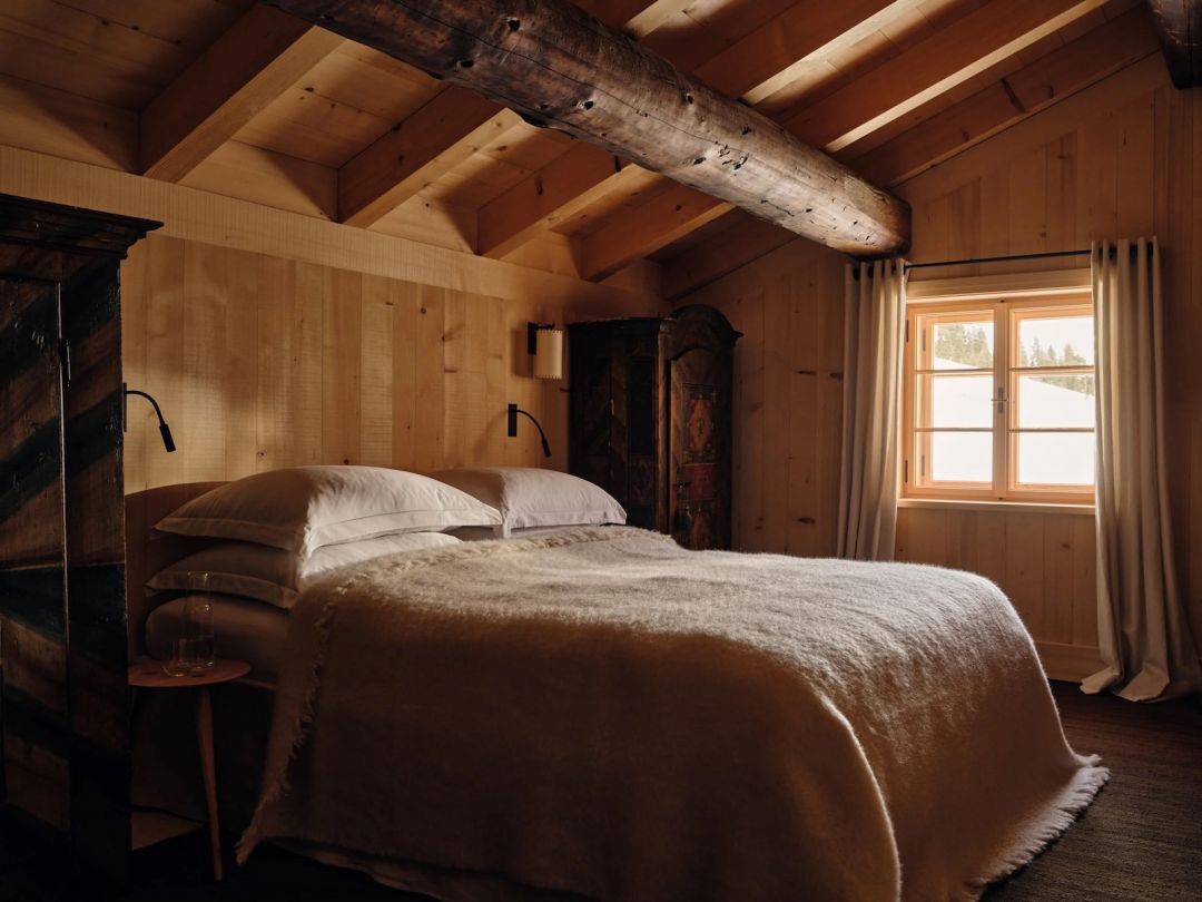 A bedroom under the roofline at the Sonnenhof, pale spruce panelling and rafters crossed by one massive old beam, white linen and a fringed wool blanket, antique painted Alpine cupboards either side.