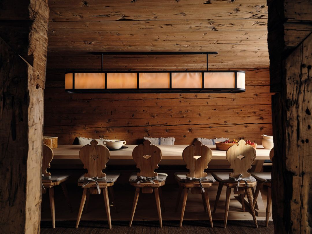 The long dining table in the Stube at the Sonnenhof, carved Alpine board chairs with heart cut-outs and tied linen cushions, a linear alabaster lantern above, framed by rough-hewn structural posts.