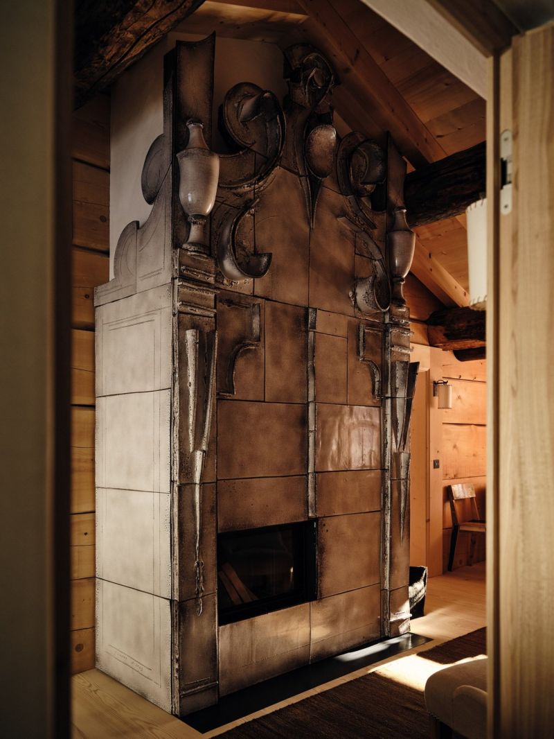 The sculptural glazed ceramic fireplace by Giuseppe Ducrot at the Sonnenhof, its bronze-toned tiles rising to modelled urns and scrolls, set against the timber wall beneath an old beam.