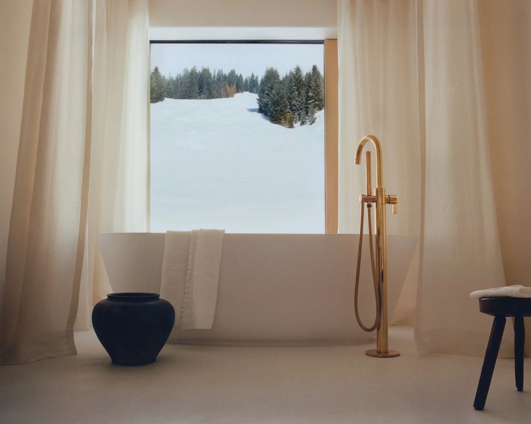 A bathroom at the Sonnenhof, a freestanding tub and floor-mounted brass tap set before a wide window onto the snow and spruce, sheer linen curtains and a black ceramic urn.