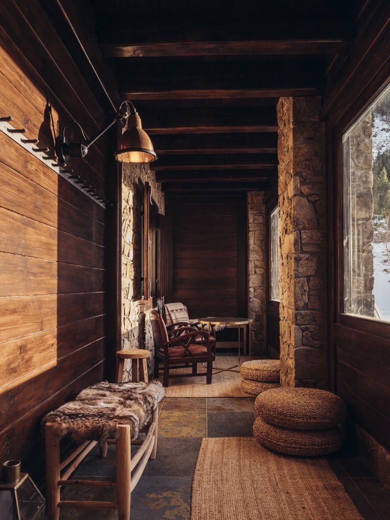 A dark timber passage at L'Ovella Negra with a brass wall lamp, stone pillar, fur-covered bench, leather armchairs and woven rattan pouffes on slate. Caption: Brass, fur and rattan against stone and dark timber.