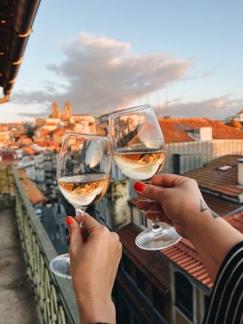 Two hands clinking white wine glasses at golden hour on a B28 Apartments balcony, looking out over Porto's old town to the silhouetted Sé Cathedral | The Aficionados 
