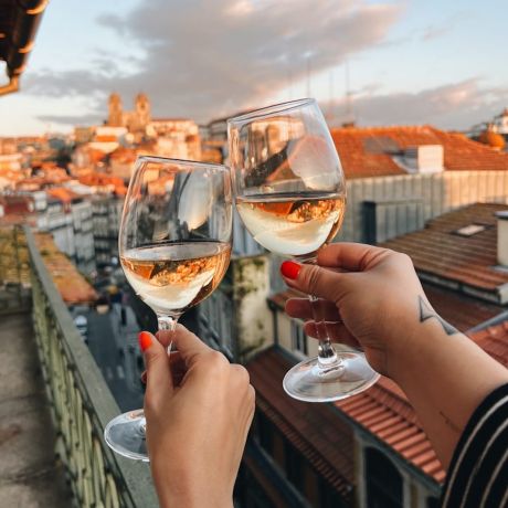 Two hands clinking white wine glasses at golden hour on a B28 Apartments balcony, looking out over Porto's old town to the silhouetted Sé Cathedral | The Aficionados 