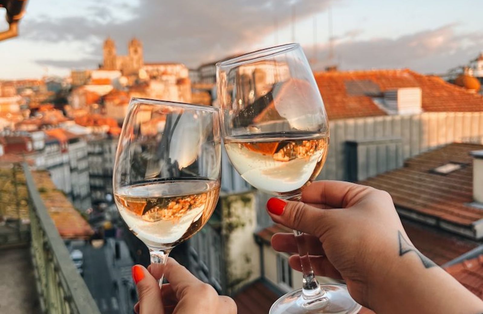 Two hands clinking white wine glasses at golden hour on a B28 Apartments balcony, looking out over Porto's old town to the silhouetted Sé Cathedral | The Aficionados 