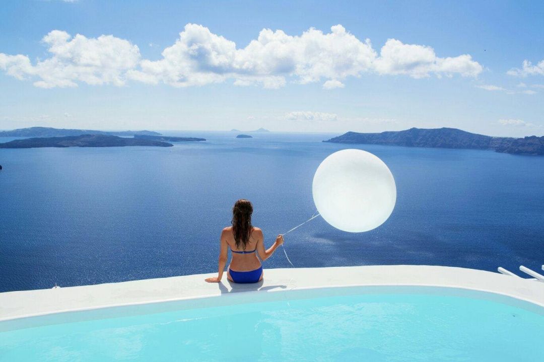 Woman sitting on the edge of an infinity pool overlooking the caldera holding a white balloon | Photo Gallery Aenaon Villas | Santorini | The Aficionados