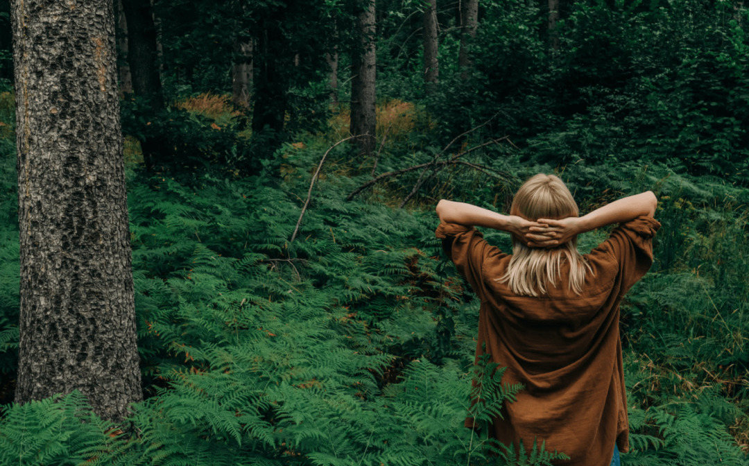 Deep forest green, Woman wearing yellow top, facing the forest holding her head in restful pose, breathing, yoga with rare oriental sweetgum trees (Liquidambar orientalis) | AHÃHA – Eco Luxury Design Hotel on Turkey’s Aegean Riviera  |The Aficionados