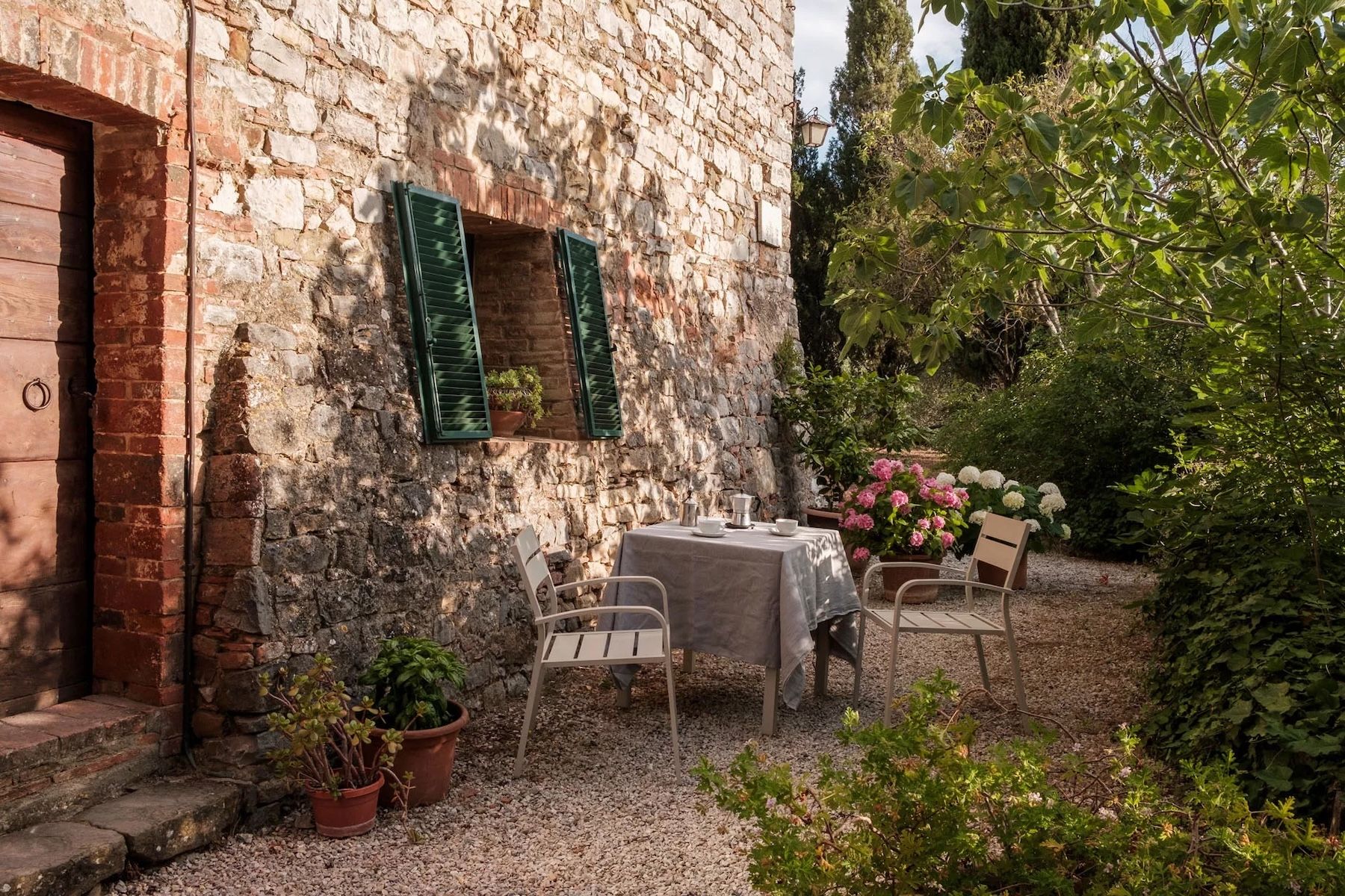 Outdoor terrace at Colle ai Lecci agriturismo, Chianti, with a linen-draped table set for coffee against a centuries-old stone and brick farmhouse facade with olive-green shutters and hydrangeas