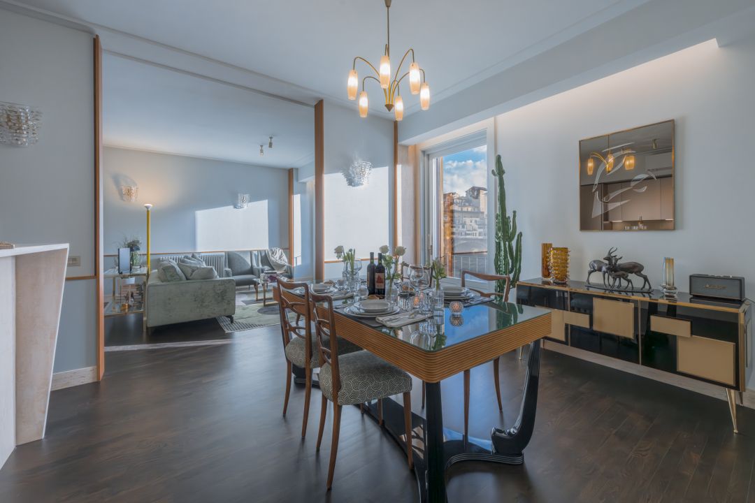 Fifties dining room at Ponte Vecchio Panorama: glass-topped table on a black lacquered base, brass chandelier, bronze deer on the sideboard and the bridge through the balcony door, images © Tommaso Pini