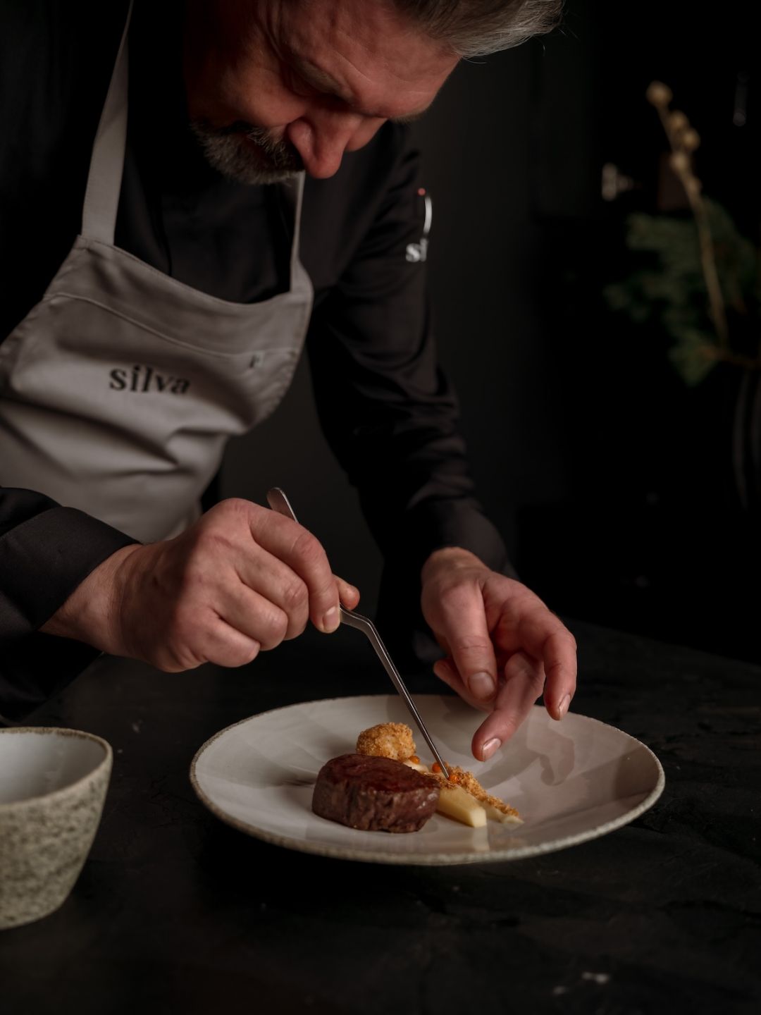 Chef Michael Helfrich in silva-branded blacks against a dark leather-tiled wall, the Noma-trained head chef of silva at Naturhotel Forsthofgut, Leogang, Austria | The Aficionados.