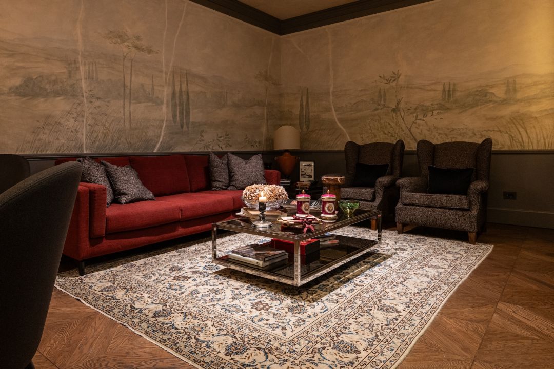 The living room at Lungarno Guicciardini 9, a brick-red sofa beneath a misty painted landscape of cypresses and hills, a glass and chrome table set with books, ribbon-tied papers and dried hydrangeas on an ivory rug. © Tommaso Pini
