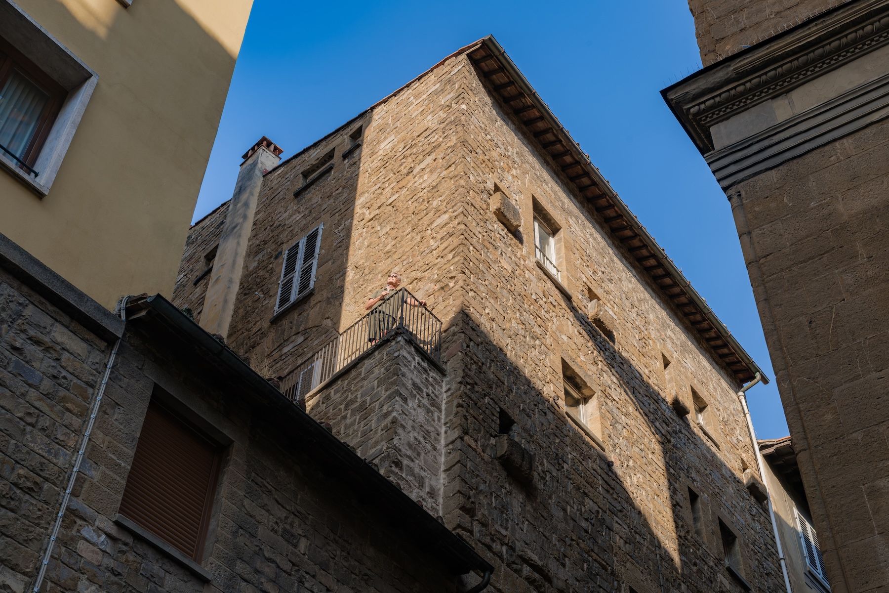 Twelfth-century stone tower-house on Via delle Terme seen from the street, a figure on its corner balcony against a clear blue sky, Terme Historical Tower, images © Tommaso Pini