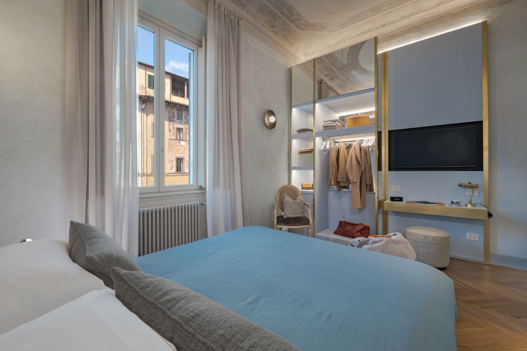 Suite 1 at Medici Historical Home: powder-blue bedcover, brass-framed wardrobe wall and a window onto a painted facade across the square, images © Tommaso Pini