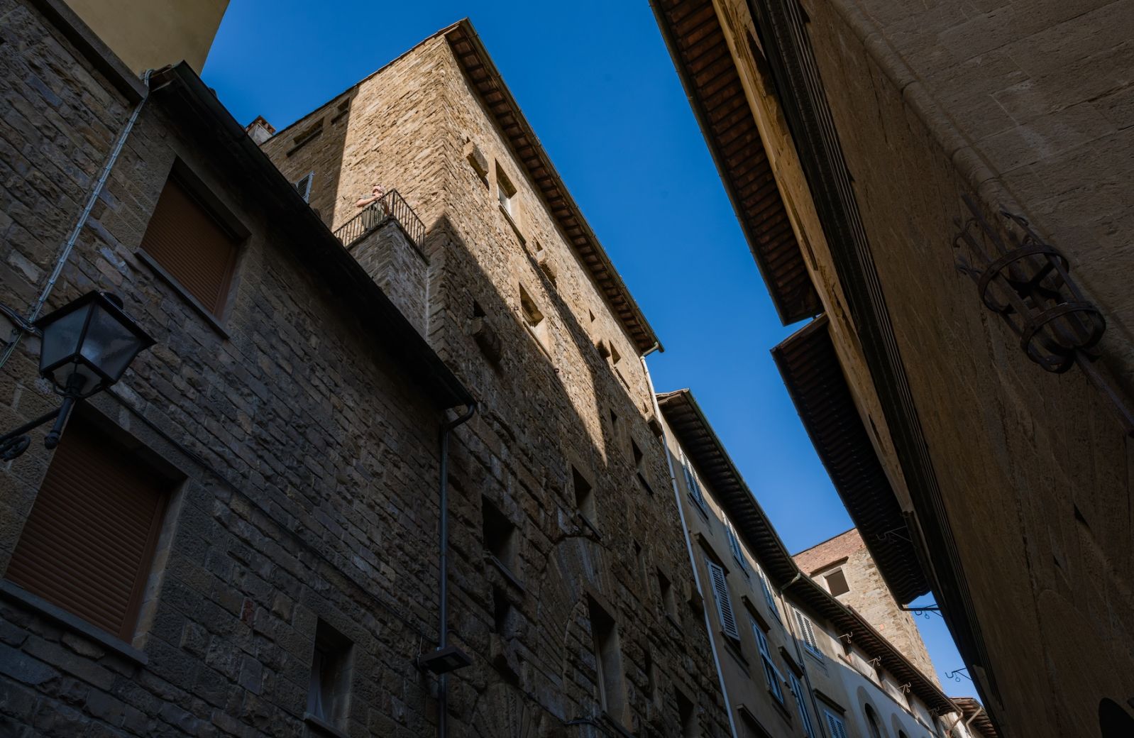 Twelfth-century stone tower-house on Via delle Terme seen from the street, a figure on its corner balcony against a clear blue sky, Terme Historical Tower, images © Tommaso Pini