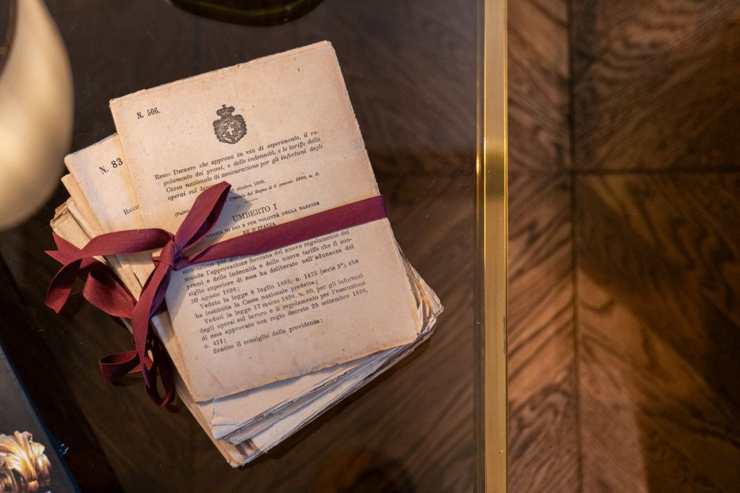 A bundle of printed decrees of the Kingdom of Italy issued under Umberto I, stamped with the Savoy arms and tied in claret ribbon, on a glass and brass table at Lungarno Guicciardini 9. © Tommaso Pini
