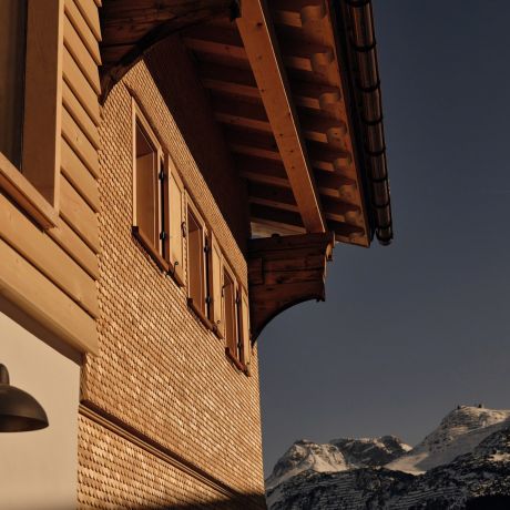 The shingled gable of Haus W in Zug at dusk, timber shutters below deep carved eaves, snow peaks beyond.