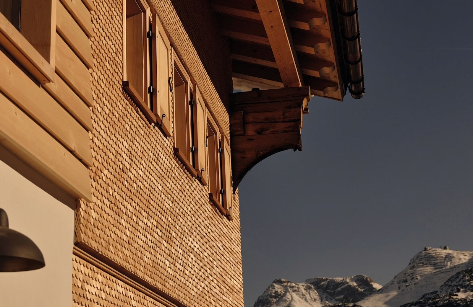 The shingled gable of Haus W in Zug at dusk, timber shutters below deep carved eaves, snow peaks beyond.