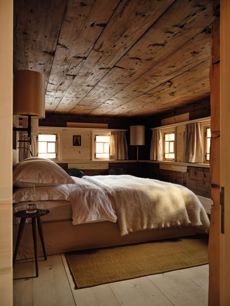 A bedroom at Haus W beneath its original 1609 ceiling of hand-split planks, small box windows set in old spruce walls, the bed in white linen under a wool blanket.