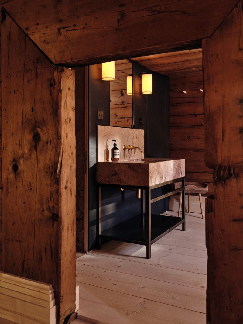 A bathroom at Haus W seen through an old timber doorway, a stone basin on a black steel stand with brass wall taps and parchment shaded lights.