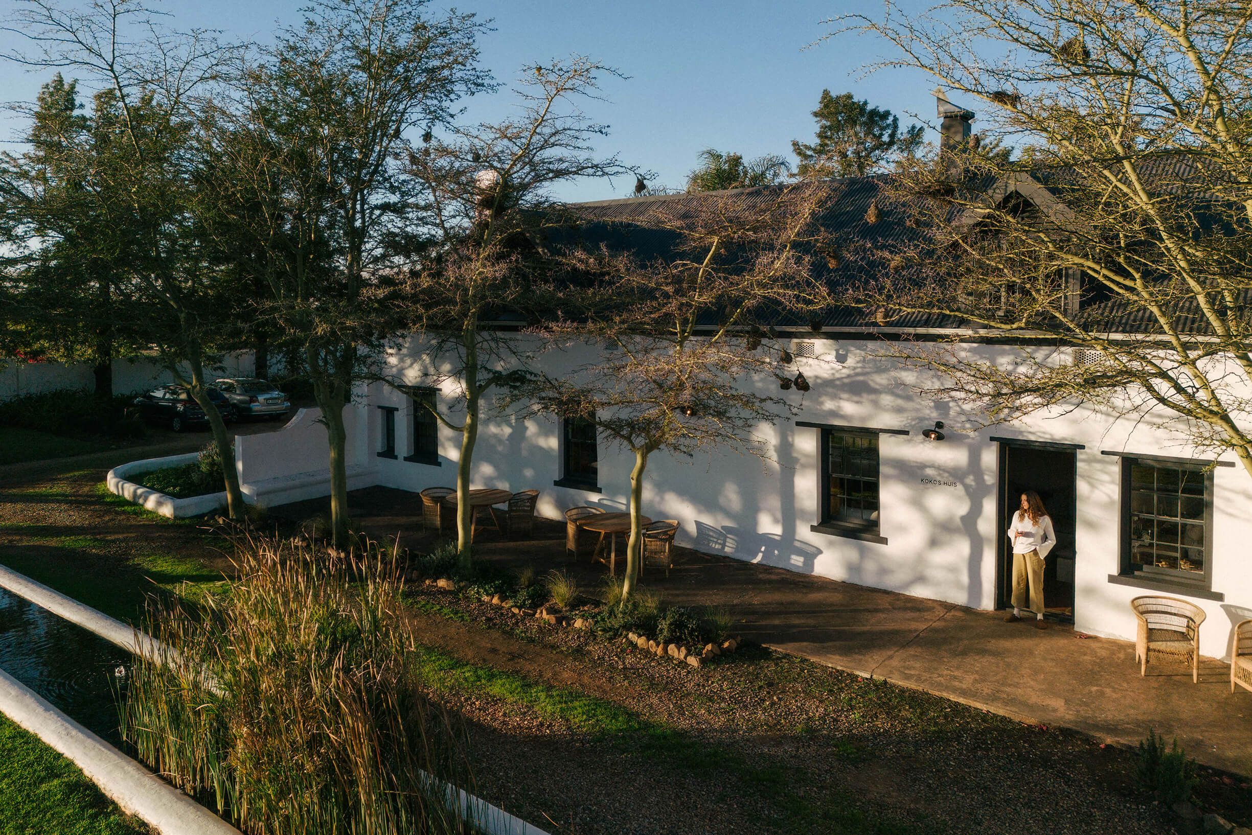 The lime-washed facade of Kokos Huis in late afternoon light, thorn trees hung with weaver nests along the stoep beneath a dark corrugated roof, a woman standing in the open doorway.