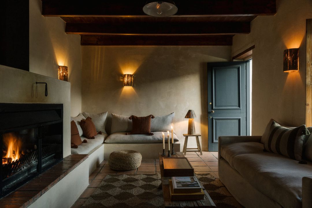 The lounge at Kokos Huis by firelight, an open black steel fireplace beside a built-in plaster banquette in linen and rust cushions, raffia sconces on buff plaster and a blue-grey panelled door.