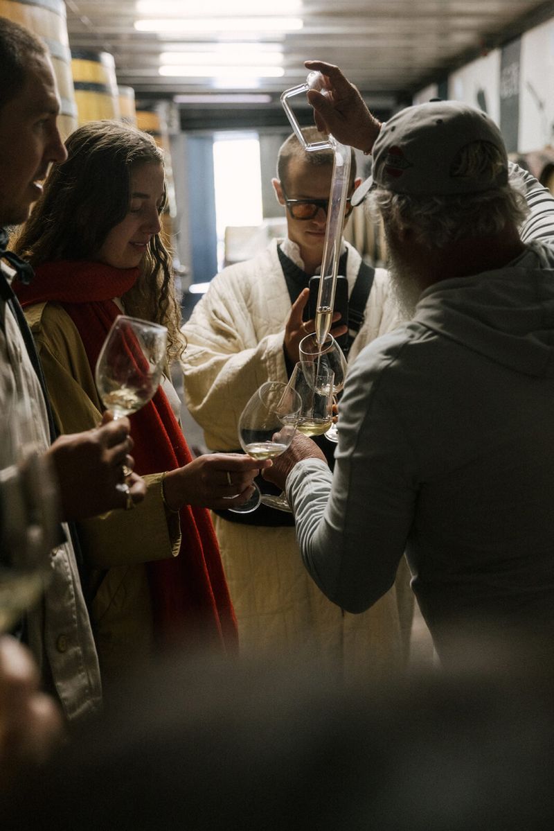A winemaker drawing wine from a barrel with a glass thief for guests holding glasses in a Swartland cellar.
