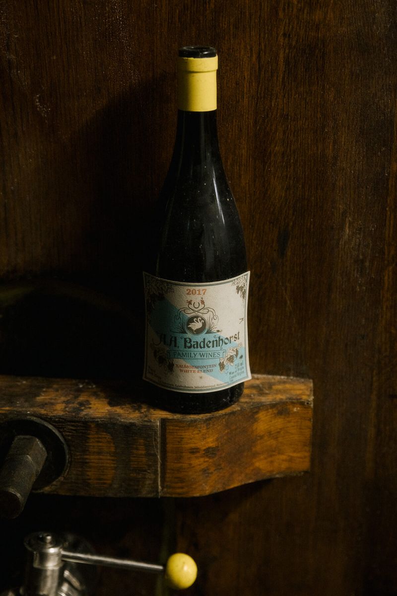 Guests tasting from the barrel with a winemaker in a Swartland cellar, alongside a bottle of A.A. Badenhorst Kalmoesfontein White Blend on a timber ledge.
