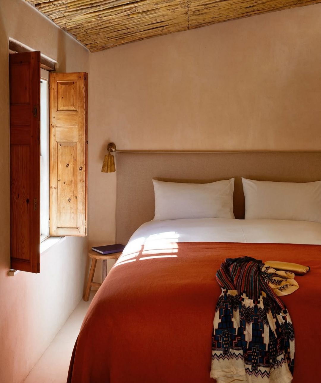 A bedroom at Kokos Huis under a split reed ceiling, morning light through an original pine shutter falling across a rust bedspread and a patterned wool throw, a brass sconce above the linen headboard.