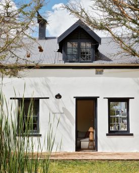 The lime-washed facade of Kokos Huis in late afternoon light, thorn trees hung with weaver nests along the stoep beneath a dark corrugated roof, a woman standing in the open doorway.