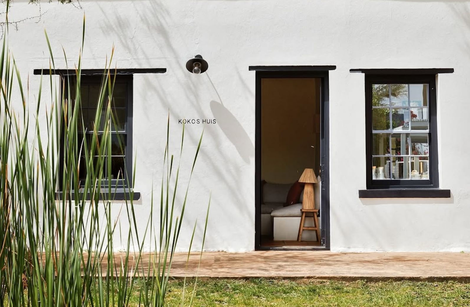 The lime-washed facade of Kokos Huis in late afternoon light, thorn trees hung with weaver nests along the stoep beneath a dark corrugated roof, a woman standing in the open doorway.