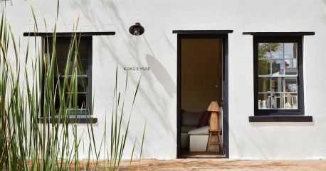 The lime-washed facade of Kokos Huis in late afternoon light, thorn trees hung with weaver nests along the stoep beneath a dark corrugated roof, a woman standing in the open doorway.