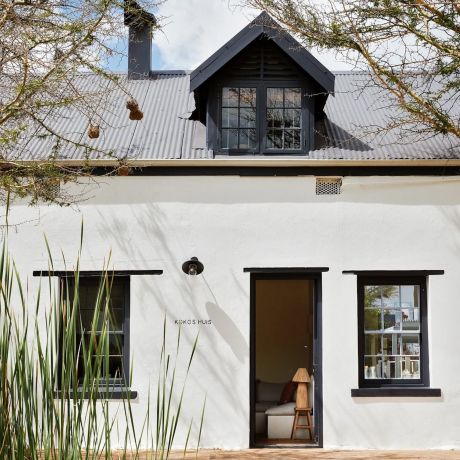 The lime-washed facade of Kokos Huis in late afternoon light, thorn trees hung with weaver nests along the stoep beneath a dark corrugated roof, a woman standing in the open doorway.