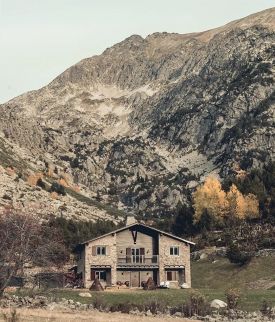 The stone and timber exterior of L'Ovella Negra in the Vall d'Incles, Andorra, with the open-sided pavilion, festoon lights and hay bales on the grass. Caption: The lodge and its pavilion at the head of the valley, hay bales still on the grass.