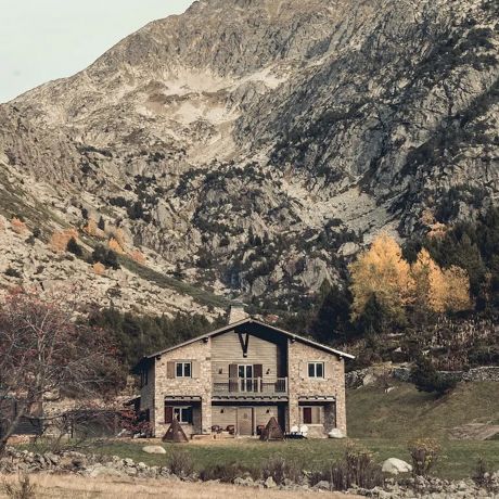 The stone and timber exterior of L'Ovella Negra in the Vall d'Incles, Andorra, with the open-sided pavilion, festoon lights and hay bales on the grass. Caption: The lodge and its pavilion at the head of the valley, hay bales still on the grass.