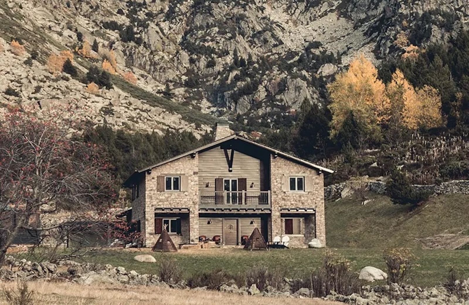 The stone and timber exterior of L'Ovella Negra in the Vall d'Incles, Andorra, with the open-sided pavilion, festoon lights and hay bales on the grass. Caption: The lodge and its pavilion at the head of the valley, hay bales still on the grass.
