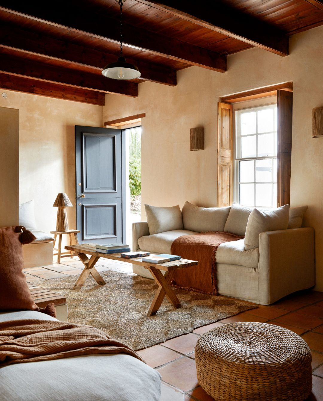 A sitting room at Kokos Huis in daylight, dark timber beams over a boarded ceiling, a linen sofa with a rust throw on old terracotta tiles, a blue-grey door open to the garden.