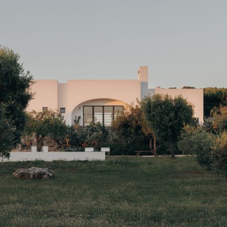 Lime-rendered facade of Masseria Moroseta caught pink in the dusk light, its arched Crittall window framed by olive trees and prickly pear, near Ostuni, Puglia | The Aficionados.