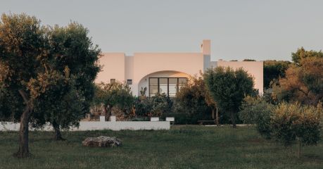 Lime-rendered facade of Masseria Moroseta caught pink in the dusk light, its arched Crittall window framed by olive trees and prickly pear, near Ostuni, Puglia | The Aficionados.