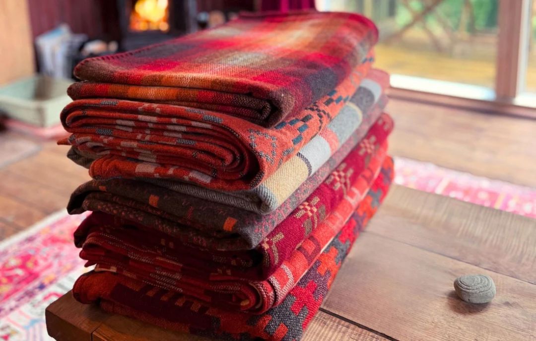 Hand Crafted Woven woolen blankets in ruby reds | Person sitting looking at the Pembrokshire coast wrapped in a blanket | Melin Tregwynt | Artisan Blankets Woven in Wales - A British Hertiage Brand | TheAficionados.com