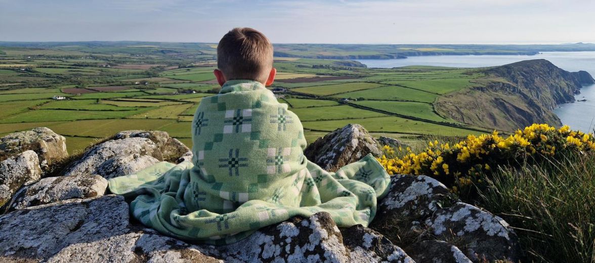 Person sitting looking at the Pembrokshire coast wrapped in a blanket | Melin Tregwynt | Artisan Blankets Woven in Wales - A British Hertiage Brand | TheAficionados.com