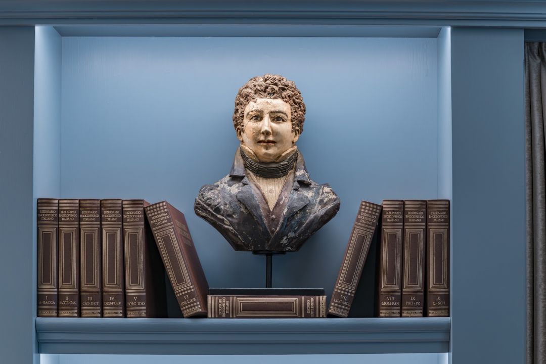 A painted nineteenth-century bust of a young man in coat and cravat, set in a carta da zucchero blue niche at Santa Croce Historical Home between volumes of an Italian encyclopaedia.