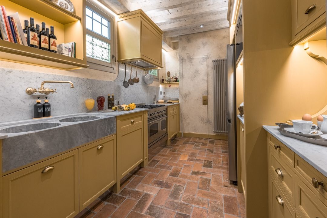 The ochre kitchen at Santa Croce Historical Home, a carved grey marble double sink, brass wall tap, Carrara splashback, range cooker and old terracotta brick floor beneath limewashed beams.