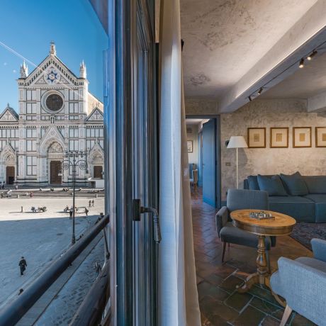 An open window at Santa Croce Historical Home framing the Basilica of Santa Croce across the piazza, beside a living room with blue sofa, walnut games table, terracotta floor and five framed drawings.