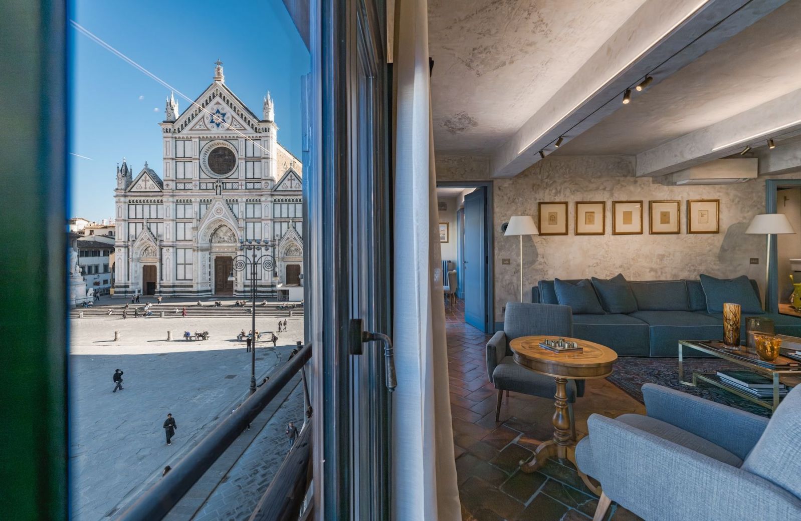An open window at Santa Croce Historical Home framing the Basilica of Santa Croce across the piazza, beside a living room with blue sofa, walnut games table, terracotta floor and five framed drawings.