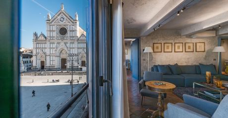 An open window at Santa Croce Historical Home framing the Basilica of Santa Croce across the piazza, beside a living room with blue sofa, walnut games table, terracotta floor and five framed drawings.